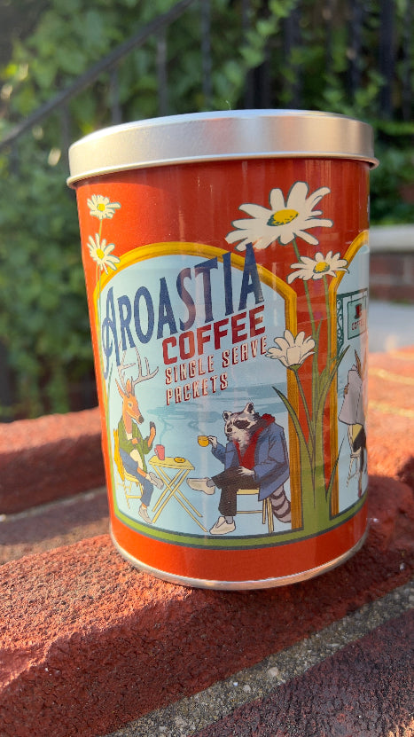 A tin container with single serve packets for coffee roasting, featuring colorful artwork on the label.