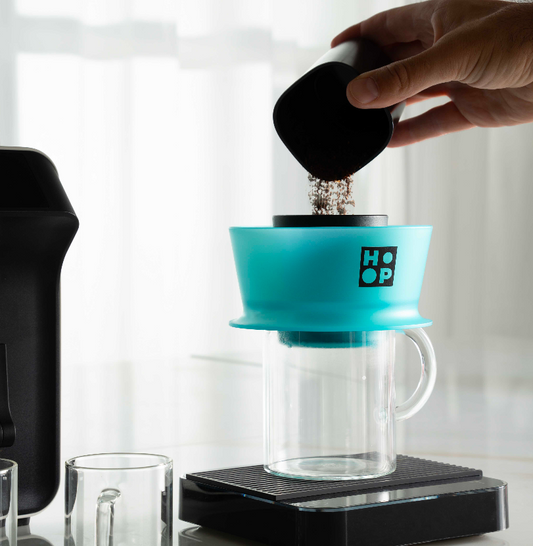 Person using a turquoise HOOP coffee maker with a clear glass carafe.